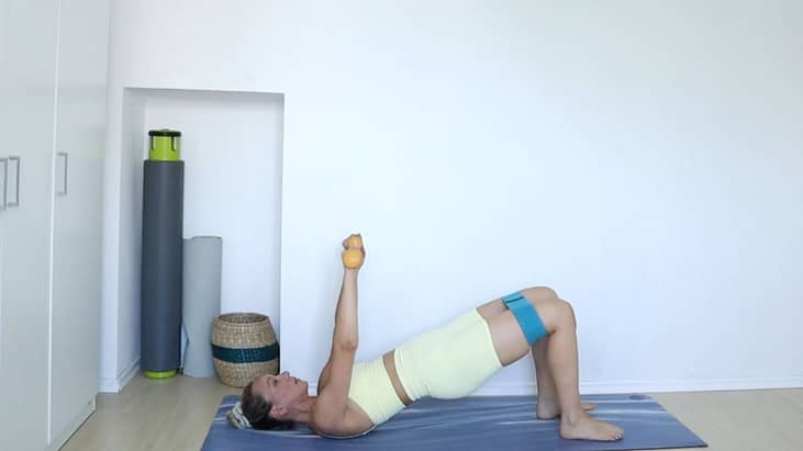 pilates intructor Ivana Devic in bridge position with arms reaching up, holding dumbbells and booty band around her thighs.