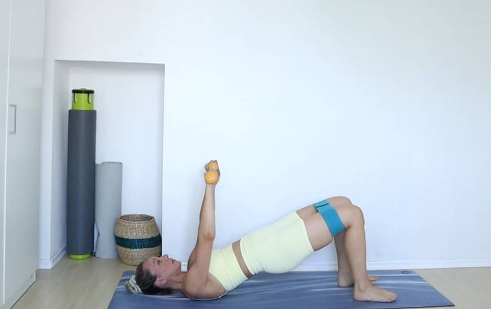 pilates intructor Ivana Devic in bridge position with arms reaching up, holding dumbbells and booty band around her thighs.