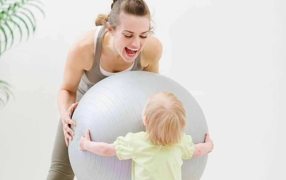 Mom wearing active wear dn playing with her toddler body by pulling a pilates ball