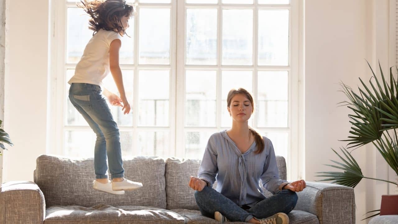 Mom is siting on the sofa and meditation while kid is jumping next to her.