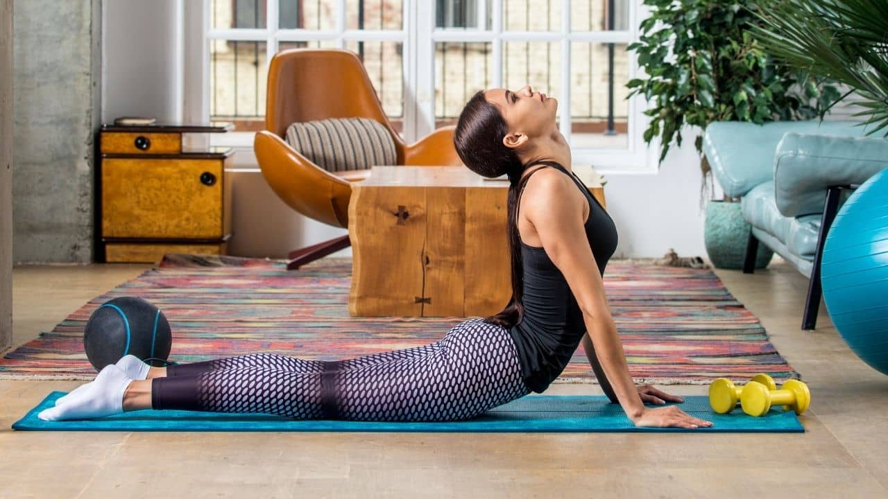 Woman in active wear, motivated for a workout, on the yoga mat, doing a Pilates swan exercise. She is surrounded by different Pilates props: dumbbells, Pilates ball, etc. She exercises in her living room.
