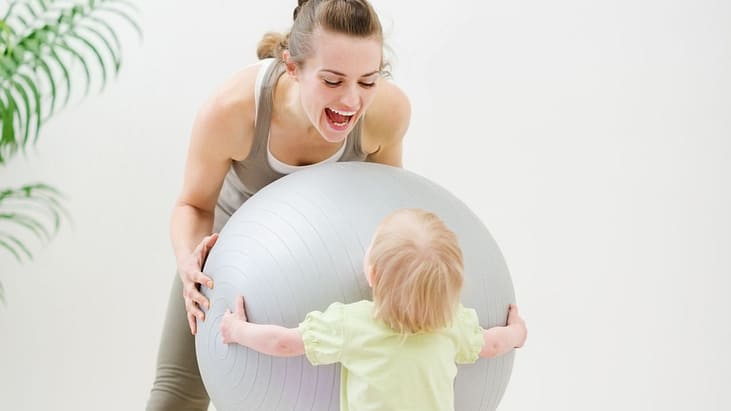 Mom wearing active wear dn playing with her toddler body by pulling a pilates ball
